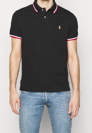 Homme portant un polo noir avec col et poignets rayés rouge et blanc, associé à un jean bleu clair, debout devant un fond blanc.