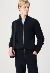 Tengerés navy ribbed zip-up pulóver magas gallérral, slim fit fazonban, elülső zsebekkel, sötét farmernadrággal párosítva.