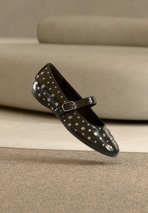 Black patent leather Mary Jane shoe with silver studs and a buckle strap, shown above a beige carpet and sofa background.