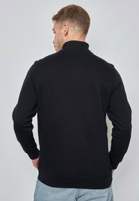 Sweatshirt noir à col montant, avec des poignets et un ourlet côtelés. Le matériau semble texturé, adapté pour un usage décontracté.