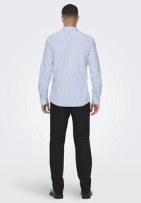 Camicia a maniche lunghe azzurro chiaro con colletto a bottone, abbinata a pantaloni neri. Materiale in cotone, texture liscia e dettagli minimi.