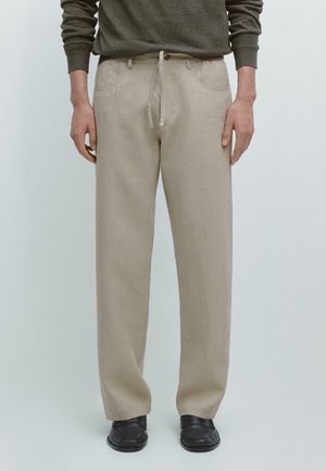 Person trägt beige Hose mit geradem Bein und Kordelzug in der Taille, dunkelgrünes Langarmshirt und schwarze Schuhe, steht vor einfachem Hintergrund.