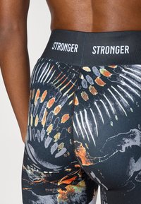 Μαύροι αθλητικοί κολάν με υφασμένο μοτίβο και πορτοκαλί και λευκές λεπτομέρειες, φαρδύ ελαστικό ζωνάρι με τη διακριτική ένδειξη "STRONGER".