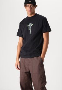 T-shirt in cotone nero con grafica di una chiave inglese verde e bianca. Abbinato a pantaloni cargo marroni, evidenziando le tasche frontali e il taglio rilassato.