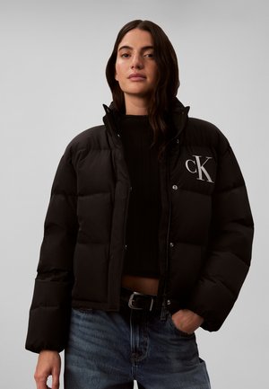 Donna che indossa una giacca imbottita nera con il logo bianco "cK" sul petto, top nero e jeans blu su sfondo grigio.