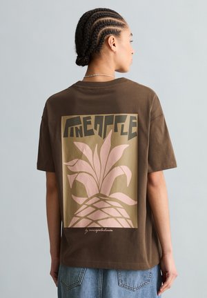 Osoba z warkoczami, ubrana w brązowy t-shirt z stylizowanym różowym motywem ananasa i napisem „Pineapple” na plecach, w połączeniu z niebieskimi dżinsami.