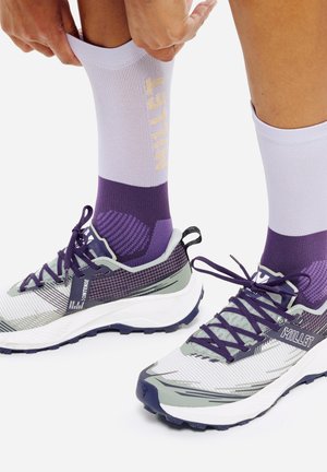 Personne en train de remonter des chaussettes violettes et blanches portées avec des chaussures de trail Millet grises à lacets violets sur fond blanc.