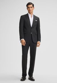 Completo nero di linea slim, con giacca a una sola bottoniera, rever a punta e due tasche frontali. Camicia bianca sotto, scarpe nere lucidate.