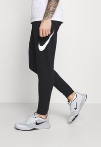 Des joggers de sport noirs présentent un logo Nike swoosh blanc, associés à des baskets grises et noires avec des détails en mesh et des accents verts.