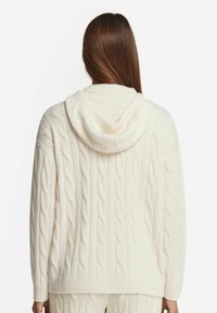 Vit kabelstickad cardigan med huva, med ribbade manschetter och nederkant. Texturen är mjuk och grov, med en avslappnad passform.