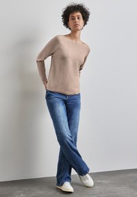 Beige Pullover mit langen Ärmeln und weitem Halsausschnitt, kombiniert mit hellblauen Schlagjeans und weißen Sneakers, vor einem neutralen Hintergrund.