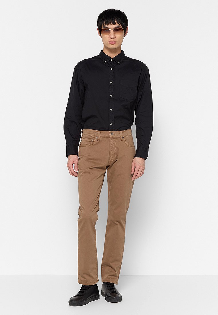 Chemise noire à boutons avec une poche poitrine, associée à un pantalon marron clair. Le modèle porte des chaussures noires et des lunettes de soleil tendance.
