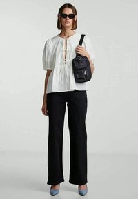 Mulher a usar uma blusa branca com atilhos, calças pretas, sapatos de salto azuis, óculos de sol e a segurar uma bolsa transversal preta.