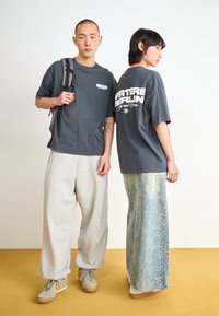 Two models wear casual outfits. One in a dark gray oversized tee and gray sweatpants, the other in a matching gray tee and snake-print wide-leg jeans.