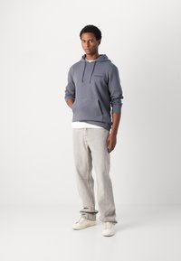 Siv hoodie s kapuco z žepom za kenguru, v kombinaciji z svetlosivimi kavbojkami in kremnimi supergami. Gladka tekstura, minimalne podrobnosti blagovne znamke.