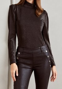 Svart, långärmad topp med hög hals och puffiga axlar i ett glänsande, texturerat tyg. Matcherande figurnära byxor med silverknappar.