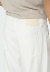 Pantalones de mezclilla blanca con dos bolsillos traseros, que cuentan con una etiqueta de cuero color marrón claro que dice "ONLY & SONS" cerca de la cintura. Textura suave.