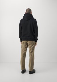 Zwarte trui met capuchon, geribbelde boorden en zoom, gecombineerd met beige corduroy broek en zwarte schoenen, gezien van achteren.