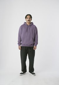 Lila Kapuzenpullover mit Fronttasche und Kordelzug, kombiniert mit schwarzen Hosen und schwarzen Sneakers mit weißen Schnürsenkeln.