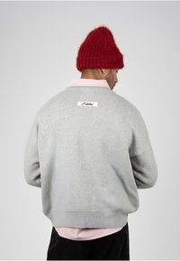Homme de dos portant un pull gris sur une chemise rose à rayures et un bonnet rouge en tricot, avec une boucle d'oreille à l'oreille gauche.
