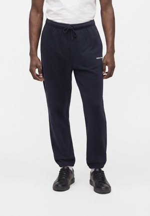 Personne portant un pantalon de jogging noir à cordon avec des bord-côtes élastiques, des baskets noires et une chemise blanche sur un fond clair uni.