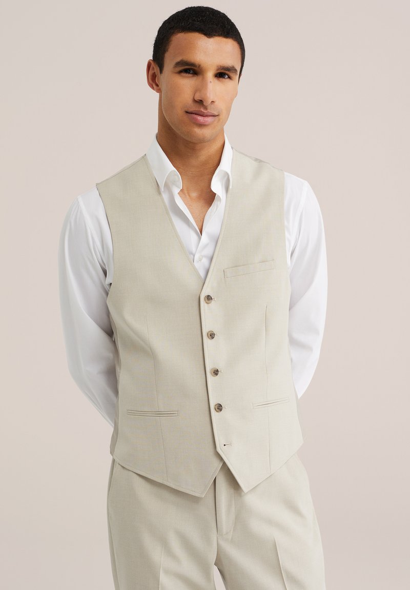 Man wearing a beige buttoned vest over a white dress shirt and matching beige trousers, standing against a plain background.
