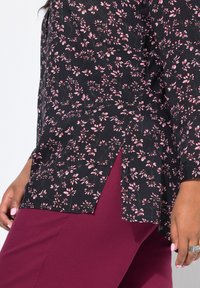 Blusa nera a fiori con fiori rosa, caratterizzata da una vestibilità comoda e spacchi laterali. Abbinata a pantaloni bordeaux scuro. Tessuto dalla texture liscia.