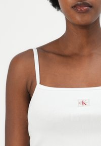 Top a canottiera bianca con coste, spalline sottili e scollatura quadrata, con un piccolo etichetta di marca rossa e beige nella zona del petto.