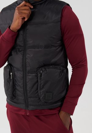 Mies, jolla on musta toppaliivi, jossa on kaksi etutaskua vetoketjulla, päällään viininpunainen pitkähihainen paita ja siihen sopivat housut, käsi lähellä kaulusta ja taskussa.