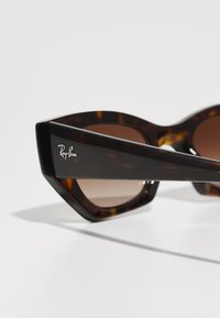 Tigrisszemezöld napszemüveg éles macska-szem formával, barna lencsékkel és Ray-Ban logóval a szárán. Simább műanyag bevonat, elegáns dizájn.
