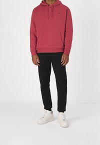 Man i röd huoodie, svarta joggerbyxor och vita sneakers står mot en enkel vit bakgrund.