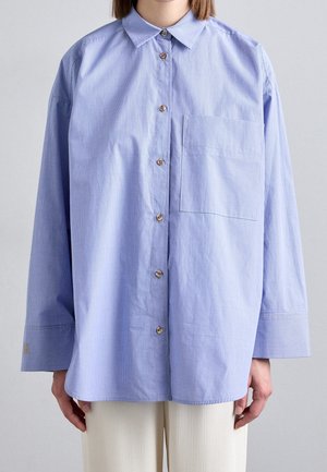 Woman wearing oversized light blue checkered button-up shirt with a chest pocket, paired with white pants, standing against a plain background.