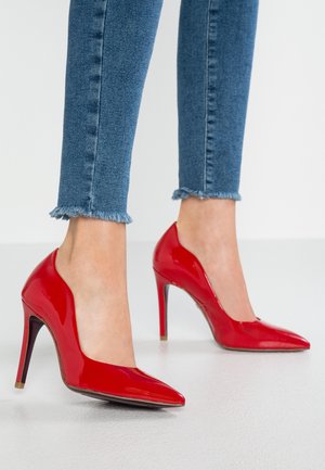 Røde laklæder høje hæle med spids tå og stiletthæl, kombineret med gennemslidte jeans med frynser ved kanten. Glinsende overflade, slankt design.