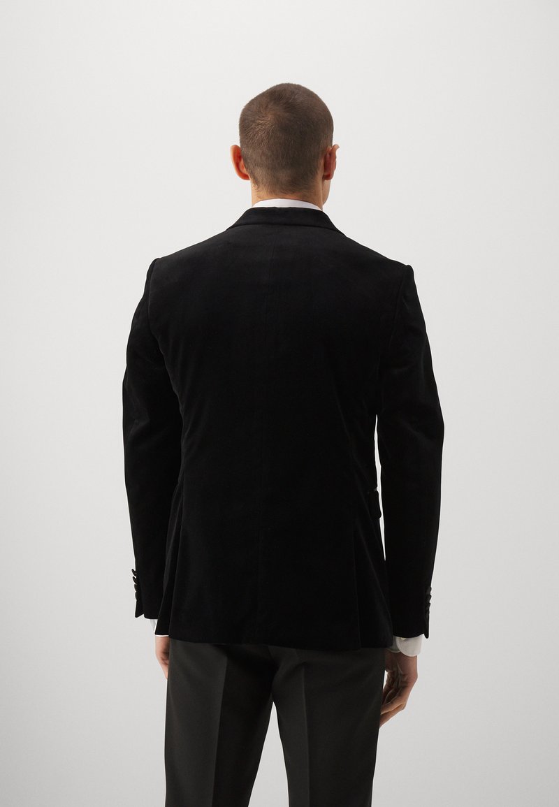 Mężczyzna z krótkimi włosami, ubrany w czarną aksamitną marynarkę oraz ciemne spodnie, stoi na jednolitym jasnoszarym tle.