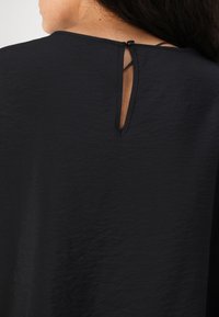 Top nero realizzato in un tessuto testurizzato con chiusura a occhiello sul retro, fissata da un piccolo bottone nero e una sottile tracolla.