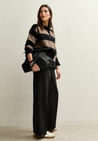 Suéter a rayas en marrón y negro con un cuello, combinado con pantalones anchos negros y zapatillas blancas. El modelo sostiene un bolso tote negro.