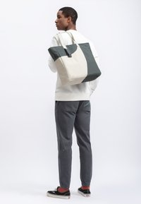 Tote bag in beige en donkergroene canvas met een tweekleurig ontwerp, stevige handvatten en een rechthoekige vorm, gedragen over de schouder van een persoon.