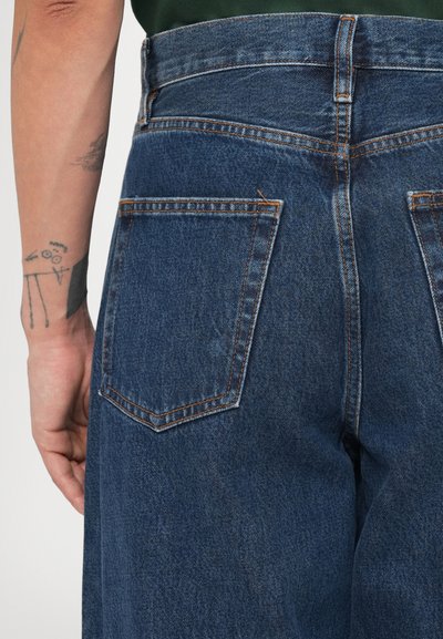 Μπλε τζιν παντελόνι με ψηλή μέση, διαθέτει δύο πίσω τσέπες και αντίθετες πορτοκαλί ραφές. Το ύφασμα έχει ελαφρώς υφή.