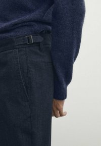 Marinblå stickad tröja med texturerad yta, som bärs över mörkgrå skräddarsydda byxor med bälte och sidoficka.