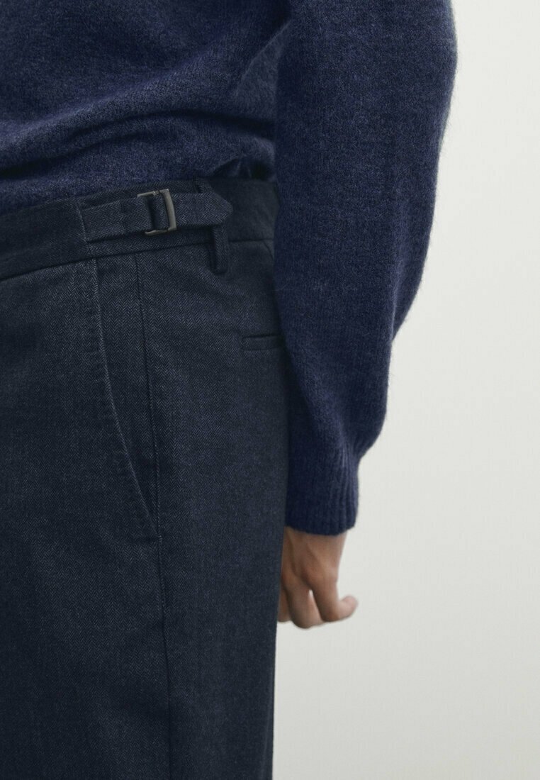 Marinblå stickad tröja med texturerad yta, som bärs över mörkgrå skräddarsydda byxor med bälte och sidoficka.