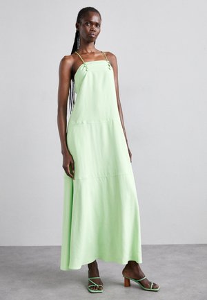 Robe maxi longue et fluide en tissu vert clair avec de fines bretelles, des liens sur les côtés et un design simple à étages. Associée à des sandales à talons fins.
