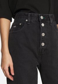 Jeans de mezclilla negros de talle alto con una bragueta de cinco botones, bolsillos laterales y un pequeño parche con el logo cerca del bolsillo; la tela tiene un acabado texturizado.