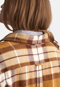 Camisa de franela de cuadros marrón y amarillo con cuello, con un forro interior a rayas y un detalle de solapa abotonada en la parte posterior.