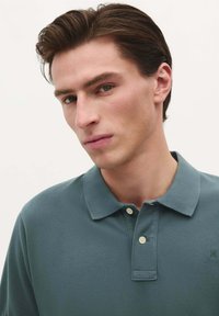 Paita polo dalam warna teal redup, terbuat dari kain katun bertekstur, dengan kerah dan placket dua kancing, serta logo di dada.