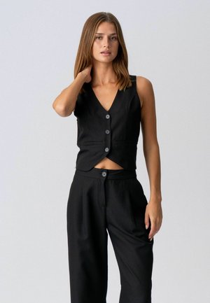 Ung kvinde med glat brunt hår iført en sort ærmeløs knapvest og sorte høj-taljede bukser, stående mod en ensfarvet baggrund.