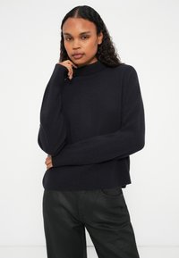 Donkerblauwe gebreide trui met een hoge kraag en een relaxte pasvorm. Heeft lange mouwen en een ribbelstructuur. Gepaard met zwarte leren broek.