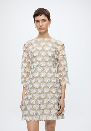 Γυναίκα με κοντά σκούρα μαλλιά που φοράει ένα μπεζ φόρεμα καλυμμένο με λευκά υφασμάτινα μοτίβα σε σχήμα βεντάλιας, στέκεται μπροστά σε ένα απλό λευκό φόντο.