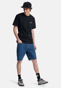 Camiseta de algodón negra, pantalones cortos de mezclilla azules, gorra negra, zapatillas grises. Presenta un logo en la camiseta y calcetines con un patrón, con las manos en los bolsillos.