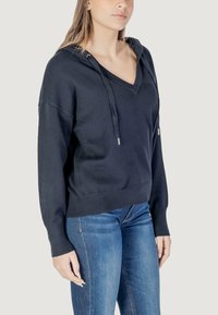 Felpa navy con scollo a V, cappuccio con lacci, polsini e orlo a costine. Tessuto morbido con una vestibilità comoda, abbinata a jeans blu.
