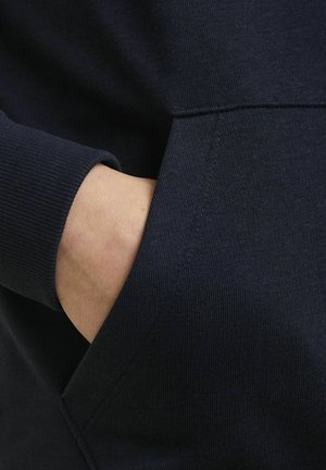 Mornarsko moder športni pulover z rebrastimi manšeti; vsebuje stransko žep, narejen iz gladkega materiala; posnetek v bližnjem načinu, na katerem je roka, ki počiva v žepu.
